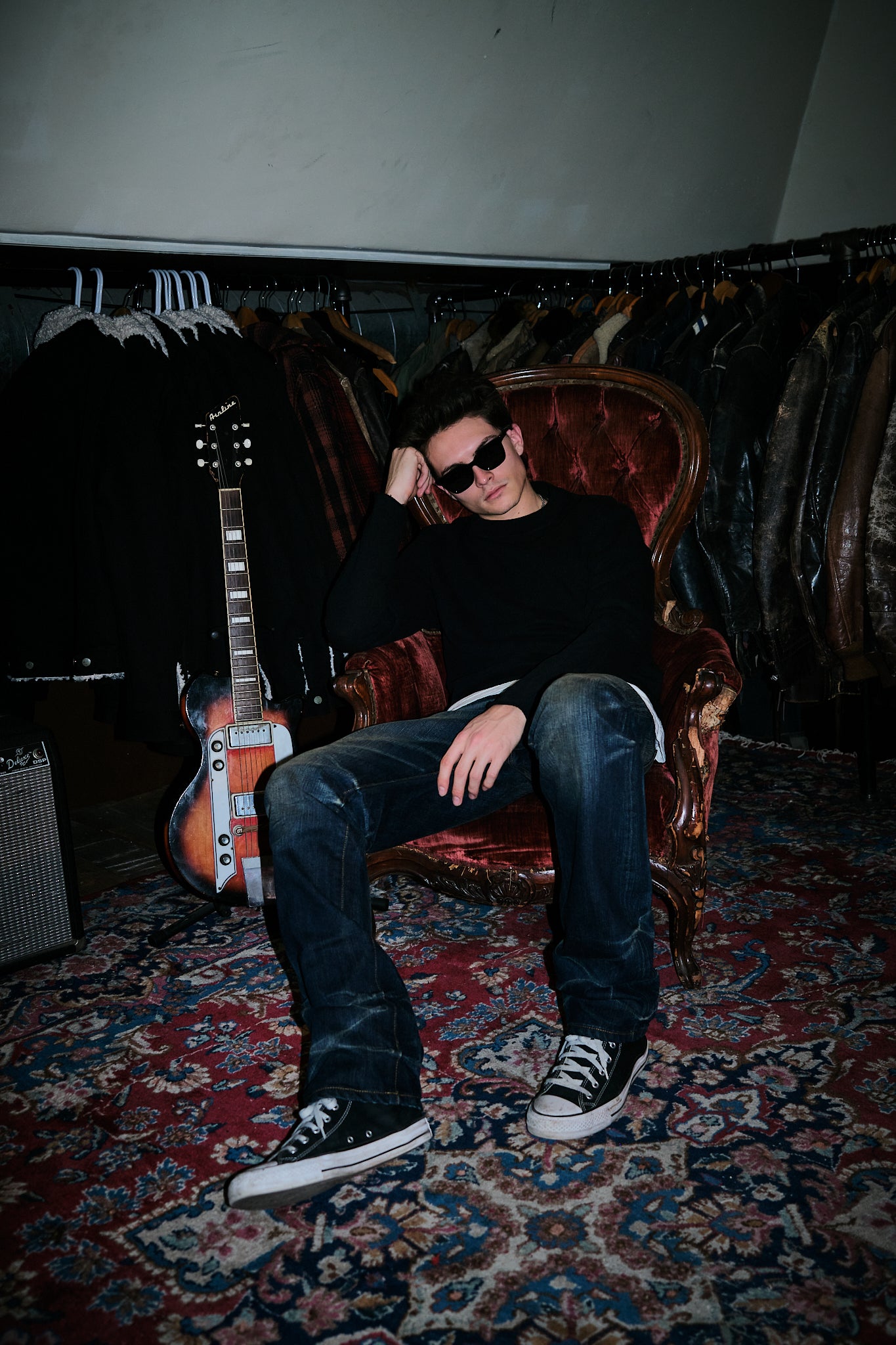 Model in sunglasses wearing the black crew sweatshirt and jeans in a velvet chair beside a guitar