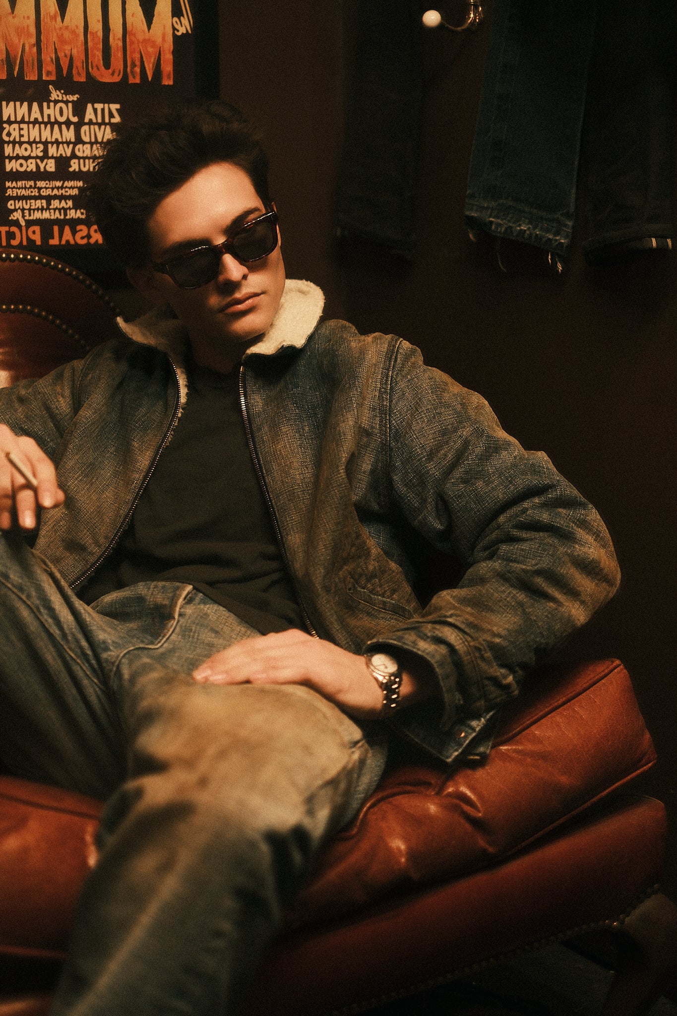 Model seated in a leather chair wearing the indigo jeans with a distressed shearling-collar jacket