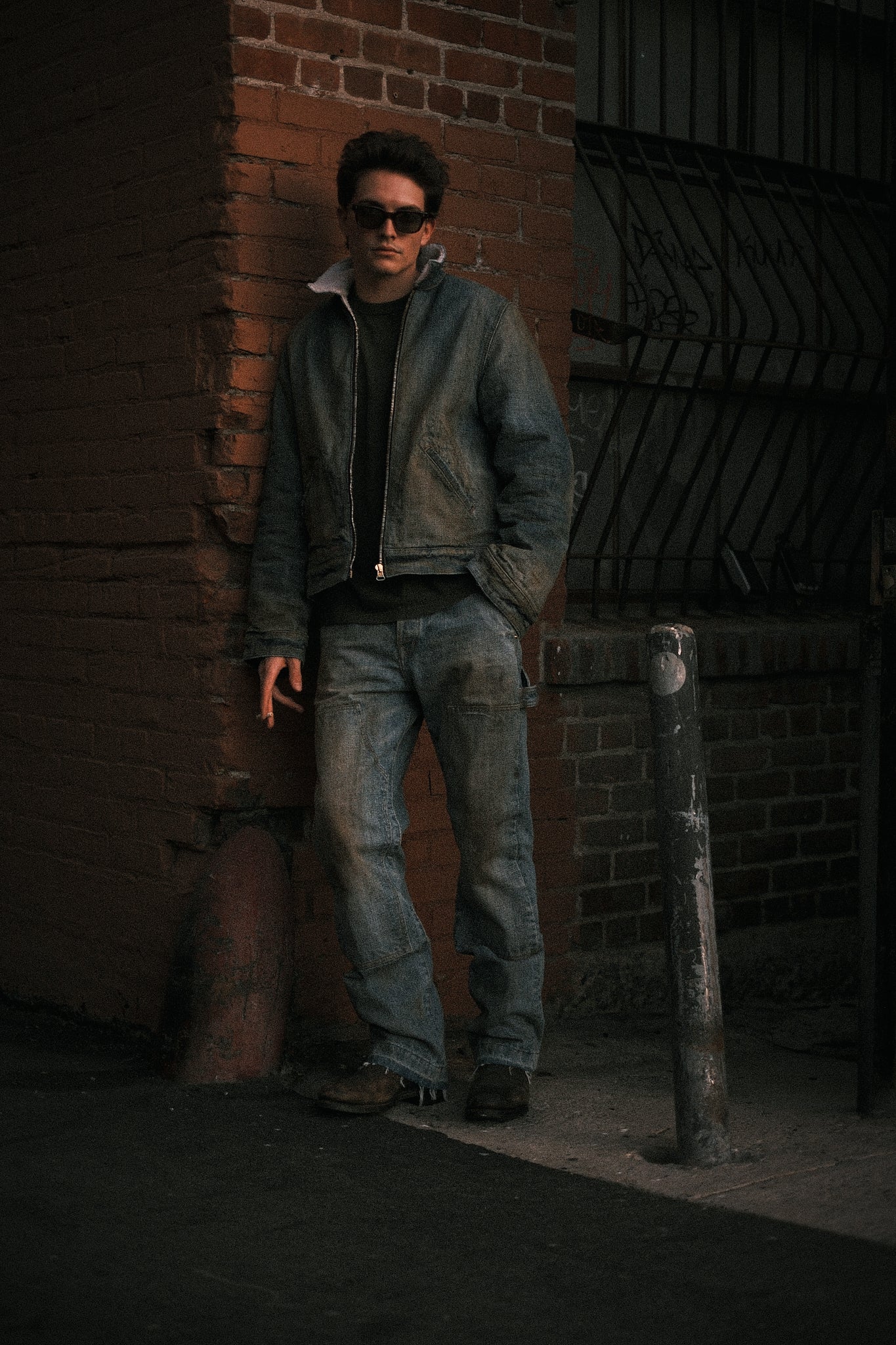 Model in sunglasses wearing the destroyed indigo jeans with a matching denim jacket by a brick wall