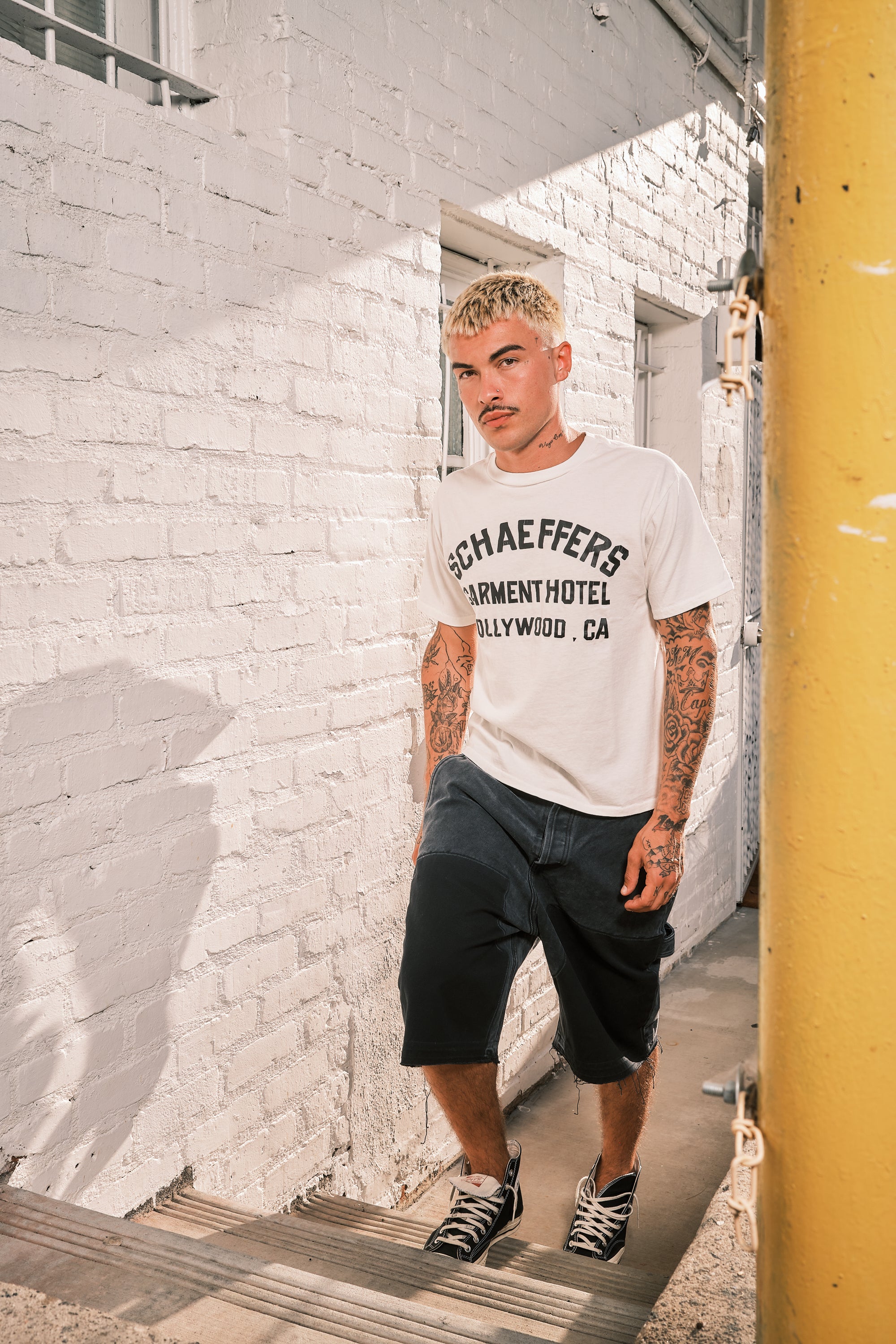 Model in a white Schaeffer's Garment Hotel tee and the black carpenter shorts leaning on a white brick wall