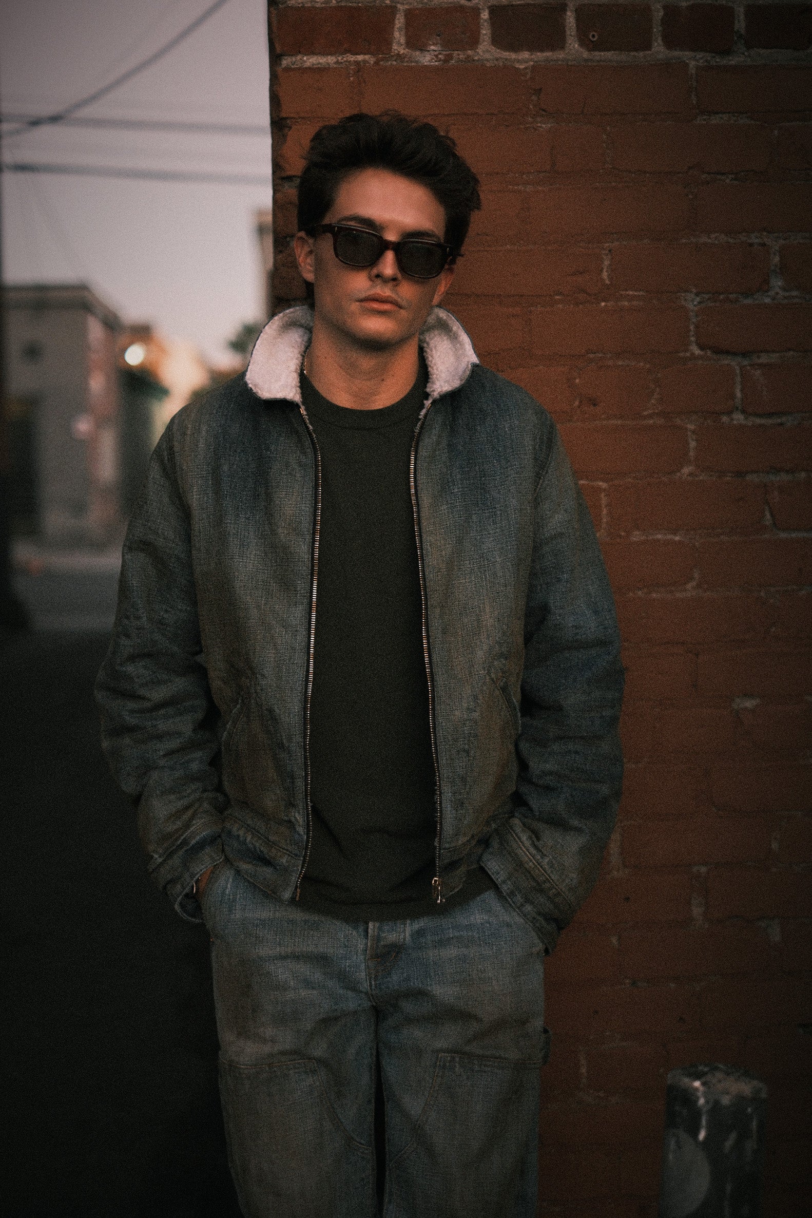 Model in sunglasses wearing the destroyed shearling denim jacket with matching jeans by a brick wall at dusk