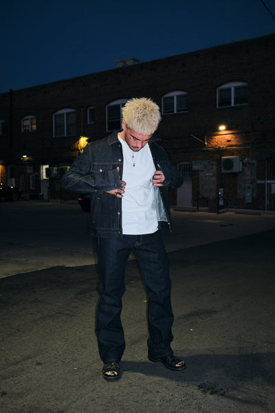 A Man Standing Out In The Open Wearing Rope indigo raw denim trucker jacket with rope indigo denim jeans with chest flap pockets and mixed metal buttons, front view