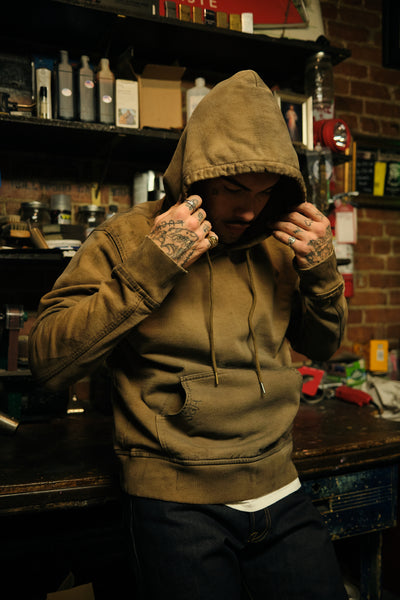 A man wearing a faded brown hooded hoodie with drawstrings, standing in a workshop or shop with shelves and a brick wall in the background.