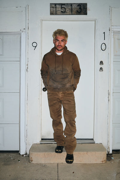 A man stands on a concrete step in front of a white door with the number 153 above it, wearing a brown hooded hoodie and matching brown jeans.